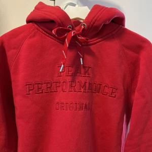 Röd hoodie från Peak Performance - Säljer en snygg och bekväm röd hoodie från Peak Performance. Den har en klassisk design med broderad logga på framsidan och en praktisk magficka. Perfekt för kyliga dagar med sin mjuka och varma känsla. Huvan har justerbara snören för extra komfort.