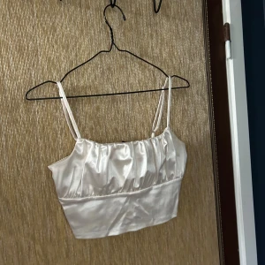 Vit satinlinne - Säljer ett elegant vitt satinlinne med smala axelband. Toppen har en glansig finish och en enkel, stilren design som passar perfekt för festliga tillfällen. Den är ärmlös och har en tight passform som framhäver figuren.