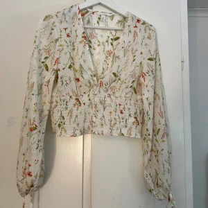 Blommig blus från H&M - Säljer en superfin blommig blus från H&M. Den är vit med ett härligt blommönster i grönt, rött och rosa. Blusen har långa ärmar och en v-ringad design med knappar framtill. Perfekt för vår och sommar! Aldrig använd!