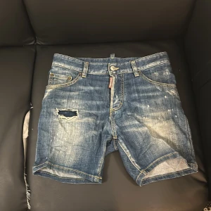 Blå jeansshorts från Dsquared2 - Säljer ett par snygga blå jeansshorts från Dsquared2. De har en cool slitning och en liten reva på framsidan som ger dem en avslappnad look. Shortsen har klassisk femficksdesign och en knappgylf. Perfekta för sommardagar!