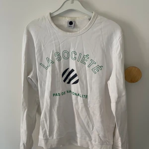 Vit långärmad tröja från La Société - Säljer en vit långärmad tröja från nn07 med texten 'LA SOCIÉTÉ' och 'PAS DE NATIONALITÉ' i grönt på framsidan. Tröjan är i bomull och har en normal passform. Perfekt för vardagsbruk eller en avslappnad dag ute. Skicka ett meddelande om du är intresserad! 😊