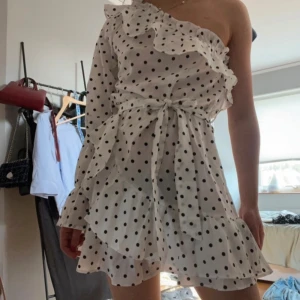 Vit prickig klänning med volanger - Säljer en superfin vit klänning med svarta prickar. Den har en oneshoulder-design och är dekorerad med volanger. Klänningen är kort och har ett knytband i midjan för en snygg passform. Perfekt för sommarens fester eller en dag på stan!