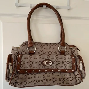 Snygg handväska - Axelväska i beige och bruna färger. Handtagen och bottnen är gjord av fakeläder. Den har två fack på sidorna och ett fack både på baksidan och inuti. Den har lite gul missfärgningar på insidan men inget som syns utanpå. 