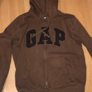 Brun hoodie från GAP - Säljer en brun hoodie från GAP i storlek S. Den har en dragkedja framtill och en stor logga på bröstet. Perfekt för höst och vinter med sin mysiga känsla. Perfekt för en avslappnad stil! Ser sprillans ny ut!