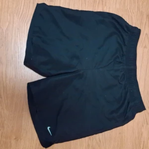 Svarta träningsshorts från Nike - Säljer ett par svarta träningsshorts från Nike. De är i bra skick och perfekta för träning eller en avslappnad dag. Shortsen har en elastisk midja för en bekväm passform. Nike-loggan syns diskret på ena benet.