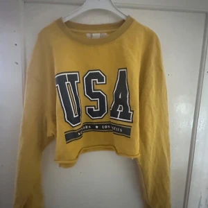 Gul croppad tröja från H&M - Säljer en cool gul croppad tröja från H&M med trycket 'USA' i stora bokstäver. Tröjan har långa ärmar och en avslappnad passform, perfekt för en casual look. Den är i bra skick och redo för nya äventyr! 🌟