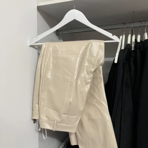 Beige byxor från Gina Tricot - Säljer ett par snygga beige byxor från Gina Tricot. De är i ett glansigt material som ger en lyxig känsla. Skinnbyxor. Byxorna har en dragkedja och knapp framtill för enkel stängning. Storlek 40, passar M. Perfekta för en stilren look! 