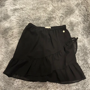 Svart kjol från Pernilla Wahlgren - Säljer en superfin svart kjol från Pernilla Wahlgren. Den har en söt volangdetalj och en omlott-design som ger en elegant touch. Storlek 38. Perfekt för både fest och vardag! 🖤