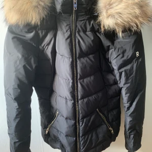 Svart dunjacka med pälskrage - Säljer en snygg svart dunjacka med en fluffig pälskrage. Jackan har en dragkedja framtill och två fickor med dragkedjor. Perfekt för kalla vinterdagar! Den är i bra skick och supervarm.