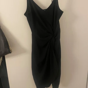 Svart klänning med knutdetalj - Elegant svart klänning med en stilren knutdetalj framtill. Klänningen är ärmlös och har en v-ringad design som ger en sofistikerad look. 