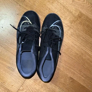 Svarta Nike fotbollsskor - Snygga svarta Nike fotbollsskor med silverdetaljer och snörning. Perfekta för inomhusspel med en slitstark sula och stilren design.
