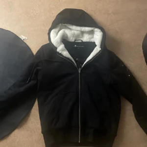 Moose Knuckles classic Bunny - Säljer en svart jacka från Moose Knuckles med dragkedja och mysigt foder. Jackan har en huva med vit fuskpäls och praktiska fickor. Perfekt för kyliga dagar.