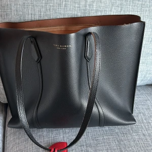 Svart shoppingväska från Tory Burch - Snygg svart shoppingväska från Tory Burch i skinn med stilren design. Väskan har långa handtag och en praktisk dragkedja upptill. Insidan är brun och rymlig, perfekt för att bära med sig allt man behöver. En röd detalj ger en fin kontrast till den svarta färgen.