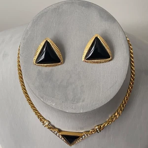 Guldfärgat vintage  smyckeset - Elegant smyckeset med ett halsband och örhängen i triangulär design. Halsbandet har en guldfärgad kedja med en svart triangulär detalj och gnistrande stenar. Örhängena matchar med samma svarta triangulära design och guldfärgad kant.