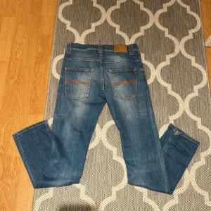 Blå jeans från Nudie Jeans - Säljer ett par snygga blå jeans från Nudie Jeans. De har en klassisk femficksdesign med orange sömmar och en knappgylf. Perfekta för en avslappnad stil. Modellen är grim tim, vilket är slimfit köpte dessa för 900 men de va för stora så jag säljer dem. Skriv under minsta fundering😊.
