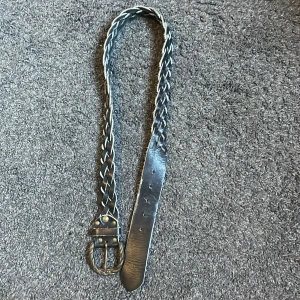 Flätat svart bälte - Flätat svart läderbälte. Bältet har en klassisk design med nitar vid spännet.   102cm långt, 3cm brett. 