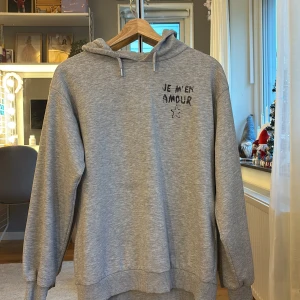 Grå hoodie från Gina Tricot - Säljer en grå hoodie från Gina Tricot med texten 'JE M'EN AMOUR' och en stjärna tryckt på framsidan. Tröjan har en bekväm passform med långa ärmar och en justerbar huva med dragsko. Perfekt för en avslappnad stil. Over sized hoodie
