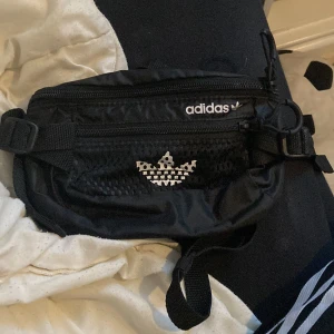 Svart midjeväska från Adidas - Snygg svart midjeväska från Adidas med justerbart bälte och dragkedja. Perfekt för att bära med sig det viktigaste. Den har ett stilrent mönster med Adidas-loggan i vitt.