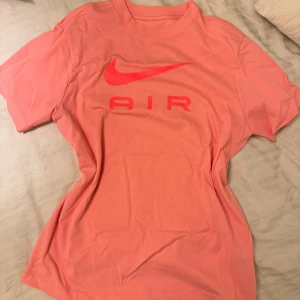Korallfärgad t-shirt från Nike - Säljer en snygg korallfärgad t-shirt från Nike med röd logga och texten 'AIR' på framsidan. T-shirten har en loose fit och är kortärmad, perfekt för en avslappnad stil.