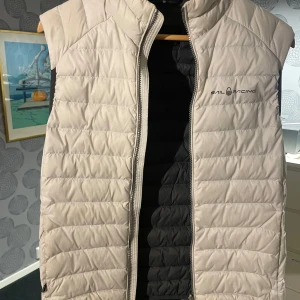 Beige quiltad väst från Sail Racing - Snygg beige quiltad väst från Sail Racing med dragkedja framtill. Perfekt för kyligare dagar och stilren design med logotyp på bröstet och ryggen.