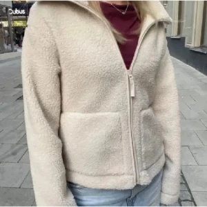 Beige/vit teddyjacka - Mysig beige/vit teddyjacka med dragkedja framtill och två praktiska fickor. Perfekt för kyliga dagar när du vill hålla dig varm och bekväm. Jackan har en klassisk krage och är långärmad. Storlek XS