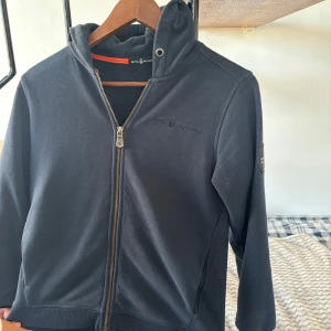 Svart huvtröja från Sail Racing - Snygg svart huvtröja från Sail Racing med dragkedja och tryck på bröstet. Tröjan har en justerbar huva och ribbade muddar. Perfekt för en avslappnad stil.