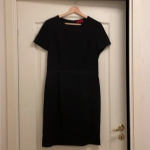 Svart klänning från Hugo Boss - Elegant svart klänning från Hugo Boss i en klassisk design. Tillverkad av 96% virgin ull och 2% elastan för en bekväm passform. Klänningen har korta ärmar och en diskret dragkedja i ryggen. Fantastiskt bra skick. Nästan ny, använd bara 1 gång. 