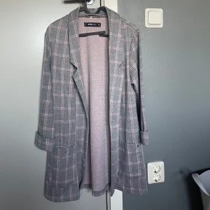 Grå rutig kappa  - Snygg grå kappa med rosa rutor. Den har en klassisk krage och långa ärmar med uppvikta muddar. Perfekt för höst och vår.