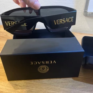 Svarta solglasögon från Versace - Snygga svarta solglasögon från Versace med breda skalmar och guldlogga på sidan. Glasögonen har en modern, rektangulär form och kommer med ett matchande svart fodral. Perfekta för en stilren look.