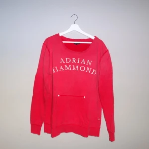 Röd sweatshirt från Adrian Hammond XL - Snygg röd sweatshirt från Adrian Hammond med tryck framtill. Tröjan har långa ärmar och en praktisk ficka på magen. Perfekt för en avslappnad stil. Storlek XL.