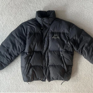 Vintage pufferjacka från Kappa - Snygg svart pufferjacka från Kappa med ikonisk logga på bröstet och ryggen. Loggan där bak funkar även som reflex. Perfekt för vintern!! Använd endast en gång!
