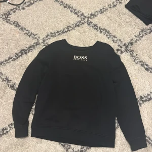 Svart tröja från Hugo Boss - Säljer en stilren svart tröja från Hugo Boss med logotyp på bröstet. Tröjan har långa ärmar och en klassisk rund halsringning. Perfekt för en avslappnad och snygg look. Lite solblekt