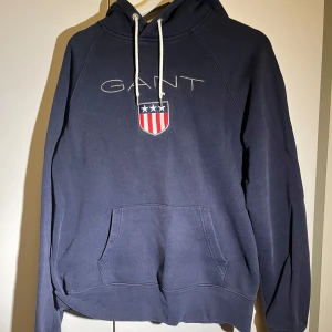 Mörkblå hoodie från GANT - Säljer en snygg mörkblå hoodie från GANT med logotyp och emblem på bröstet. Tröjan har en klassisk känguruficka och justerbar huva med vita snören. Perfekt för en avslappnad stil.