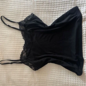 Svart linne med spetsdetaljer från Bik Bok - Snyggt svart linne från Bik Bok med tunna axelband och vacker spetsdetalj vid halsringningen. Perfekt för en stilren look.