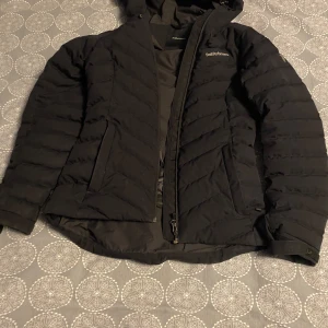 Svart dunjacka från Peak Performance - Snygg svart dunjacka från Peak Performance med dragkedja och huva. Perfekt för kyliga dagar med sin quiltade design och varma fyllning. Jackan har en stilren look med diskret logga på bröstet och ärmen. Inga fläckar, dåligt ljus på bilderna. Be om fler bilder så skickar jag. 