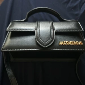 Svart handväska från Jacquemus - Elegant svart handväska från Jacquemus i skinn med ett stilrent och kompakt design. Väskan har ett handtag upptill och en axelrem för mångsidig användning. Den har en guldlogotyp på framsidan och en diskret ficka på baksidan.