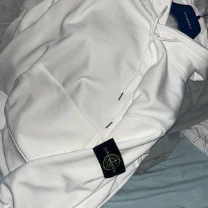 Stone Island hoodie  - Säljer en stilren vit hoodie från Stone Island med klassisk patch på ärmen. Hoodien har en stor ficka framtill och justerbar dragsko i huvan. Perfekt för en avslappnad look.