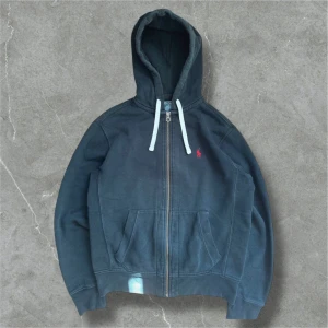 Svart Hoodie från Ralph Lauren - Säljer en snygg svart hoodie från Polo Ralph Lauren med dragkedja och vit snörning i huvan. Den har en liten röd logga på bröstet och praktiska fickor framtill. Perfekt för en avslappnad stil.