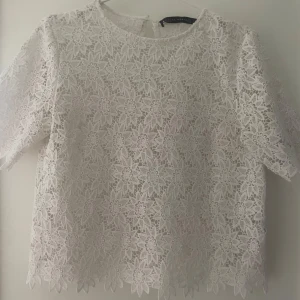 Vit spetsblus från Zara - Säljer en elegant vit spetsblus från Zara med korta ärmar och ett vackert blommönster. Perfekt för en stilren look. Blusen har en rund hals och är lätt att matcha med olika outfits.