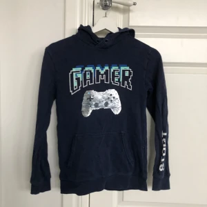 Mörkblå hoodie med gamer-tryck från H&M - Cool mörkblå hoodie från H&M med ett stort gamer-tryck och en spelkontroll i paljetter på framsidan. Tröjan har långa ärmar och en praktisk ficka framtill. Perfekt för gaming-entusiaster!
