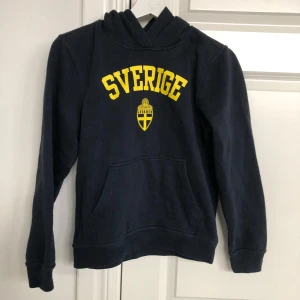 Mörkblå hoodie från Clique med Sverige-tryck - Säljer en mörkblå hoodie från Clique med gult Sverige-tryck och emblem på framsidan. Tröjan har en klassisk känguruficka och långa ärmar. Perfekt för att visa ditt stöd för Sverige!