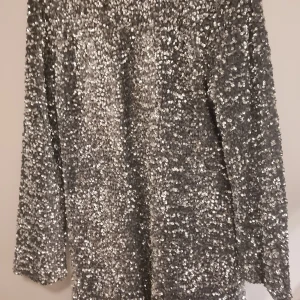 Silvrig paljettklänning med öppen rygg - Snygg och glittrig klänning i silver med paljetter över hela ytan. Den har en öppen rygg och långa ärmar, perfekt för en festlig kväll. Klänningen är i one size och ger en glamorös look. Helt oanvänd.  