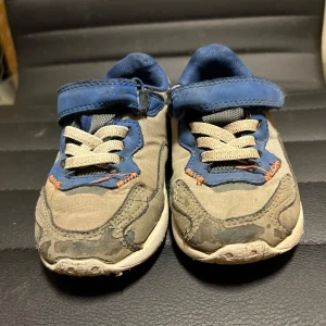 Grå och blå sneakers med kardborreband - Säljer ett par grå sneakers med blå detaljer och kardborreband. Skorna har snörning och en bekväm sula, perfekta för vardagsbruk.