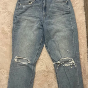 Blå jeans från American Eagle - Säljer ett par blå jeans från American Eagle med slitna detaljer på knäna. De har en klassisk femficksdesign och knappgylf. Perfekta för en avslappnad stil.