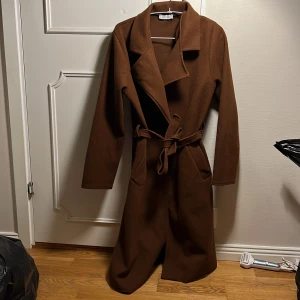 Brun kappa från Cosy House - Snygg brun kappa från Cosy House med knytbälte i midjan. Perfekt för höst och vår. Långärmad design med klassisk krage och stilren look. Ett måste för garderoben!