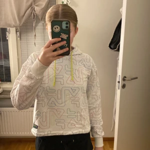 Vit hoodie från FILA med färgglatt mönster - Säljer en vit hoodie från FILA med ett färgglatt, abstrakt mönster. Hoodien har en justerbar huva med neon gröna snörrre och en avslappnad passform. Perfekt för en casual look!