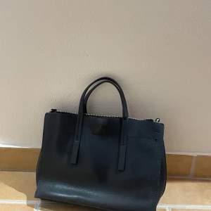 Svart handväska i skinn - Elegant svart handväska i skinn med stilrena handtag och dragkedja upptill. Den är i super bra skick 