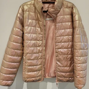 Guldglänsande pufferjacka från Active Wear - Snygg och trendig guldglänsande pufferjacka från Active Wear. Jackan har en dragkedja framtill och är perfekt för kyligare dagar. Den quiltade designen ger en modern look och den är lätt att matcha med olika outfits.