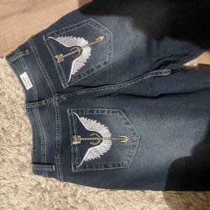 Mörkblå jeans med broderade fickor - Snygga mörkblå jeans med unika broderade detaljer på bakfickorna. Byxorna har en klassisk bootcut-stil och är perfekta för en avslappnad look. De är tillverkade i ett slitstarkt denimtyg och har en bekväm passform.
