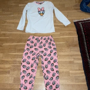Rosa och vitt piamas/mjukis sett med Daisy-mönster - Säljer ett par rosa pyjamas/mjukis byxor med Daisy Duck mönster och en vit piamas tröja med Daisy Duck på.  Byxorna har en elastisk midja och muddar vid bensluten för extra komfort. Perfekt för både Disney fans och sina som bara vill ha en mysig hemmakväll!!!